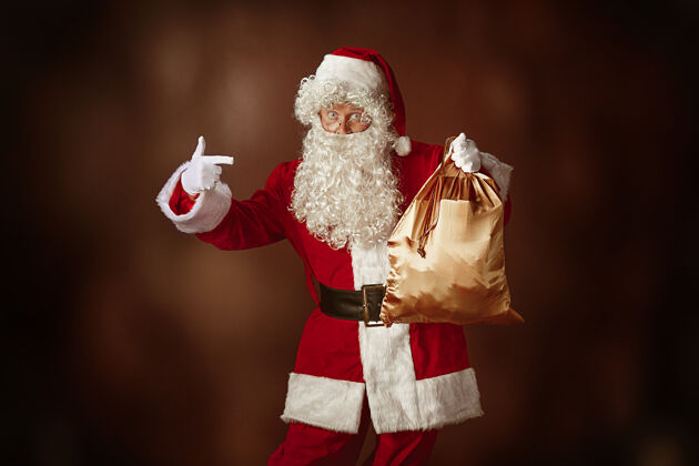 圣诞节穿着圣诞老人服装的人的肖像-一个豪华的白胡子 圣诞老人的帽子和红色服装在红色工作室的背景与礼物礼物人圣人