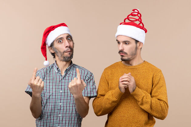 看正面图两个戴着圣诞帽的感兴趣的男人一个看着另一个在米色孤立的背景上帽子男人圣诞老人