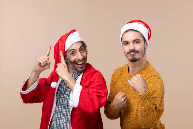 展示正面图两个戴着圣诞帽的朋友一个手指在上面露出什么东西 另一个手指在米色孤立的背景上露出拳头圣诞老人观点帽子