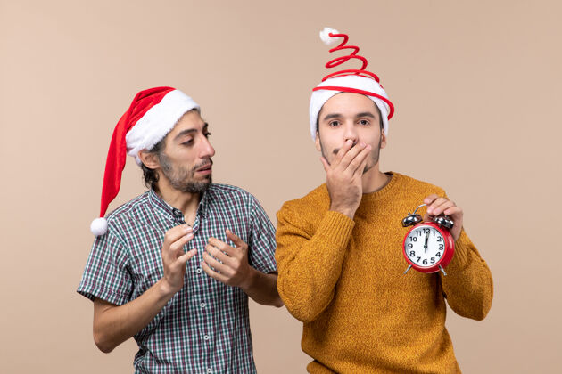 手前视图两个圣诞节男人一个拿着闹钟 用手捂住嘴 背景是米色的男人帽子男人