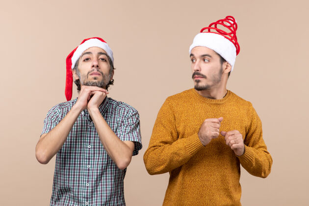 下巴前视图两个戴着圣诞帽的家伙一个把手放在下巴上 另一个在米色孤立背景下看着他两个伙计们肖像两个