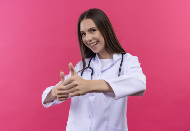 医生微笑的年轻医生女孩穿着听诊器医用长袍 在孤立的粉色背景上竖起大拇指穿着粉红医疗