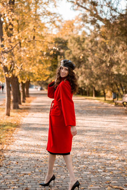 苗条身材高挑迷人时尚微笑瘦削的女人留着卷发漫步在公园里穿着温暖的红色外套秋日时髦 街头风十足 戴着贝雷帽时尚连衣裙服装