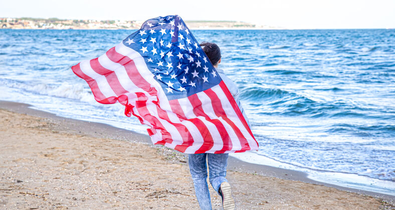 文化一位手持美国国旗的年轻女子在海边奔跑爱国主义和独立日庆祝活动的概念夏天美国爱国者