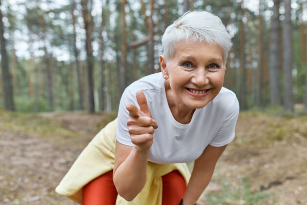户外户外风景充满活力的快乐的女退休人员在运动服前俯身 微笑着 手指在前面运动休息人