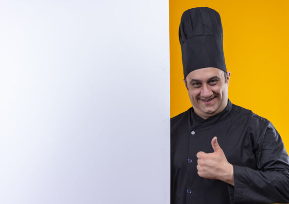 男性微笑的中年男厨师穿着厨师制服 举着白色的墙壁 他的大拇指放在黄色的背景上 留着复印空间手持向上拇指