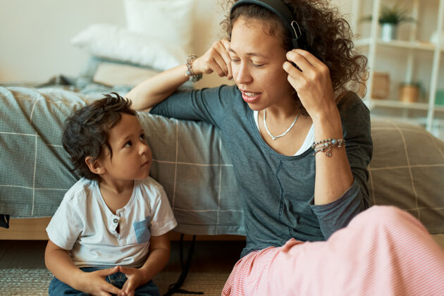 坐着美丽的西班牙年轻女子坐在地板上 戴着无线耳机和她英俊的小儿子一起听音乐的横向肖像快乐的家庭在家里玩得开心家长种族耳机