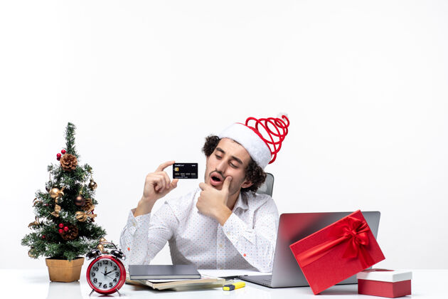 圣诞老人年轻忙碌的商务人士戴着圣诞老人的帽子 拿着他的银行卡 在白色背景的办公室里仔细地头脑风暴办公室商务人士人