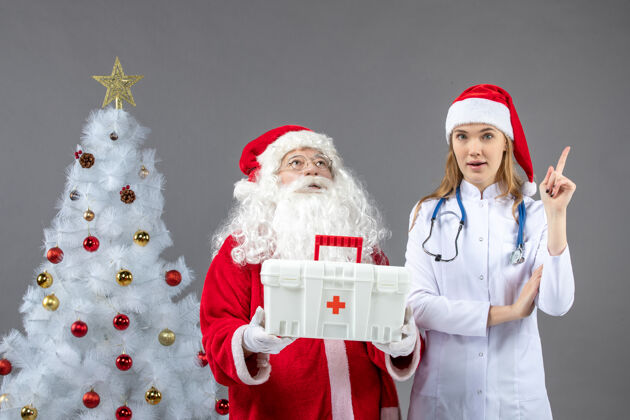 节日圣诞老人和给他急救箱的女医生在灰色墙上的正视图圣诞健康快乐