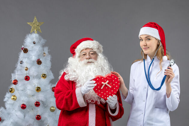 人圣诞老人和女医生的前视图 女医生在灰色墙上用听诊器观察礼物圣诞老人快乐人们