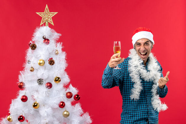 年轻人圣诞节心情与情绪疯狂的年轻人与圣诞树附近拿着一杯葡萄酒的蓝色条纹衬衫圣诞老人帽子举行聚会小伙子