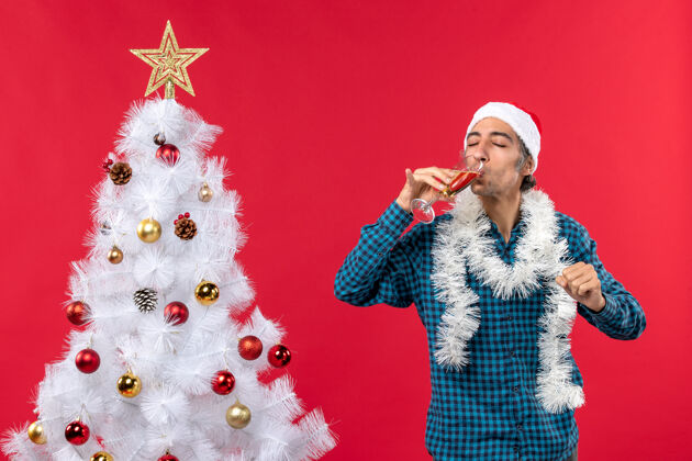 喝酒圣诞节心情与情绪激动的年轻人与圣诞老人帽子在一个蓝色的剥夺衬衫举行和喝一杯葡萄酒闭上眼睛靠近圣诞树男人圣诞树年