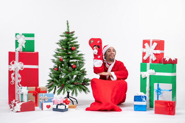 季节性微笑的圣诞老人坐在地上 穿着圣诞袜 靠近礼物和装饰在白色背景上的新年树地圣诞老人袜子
