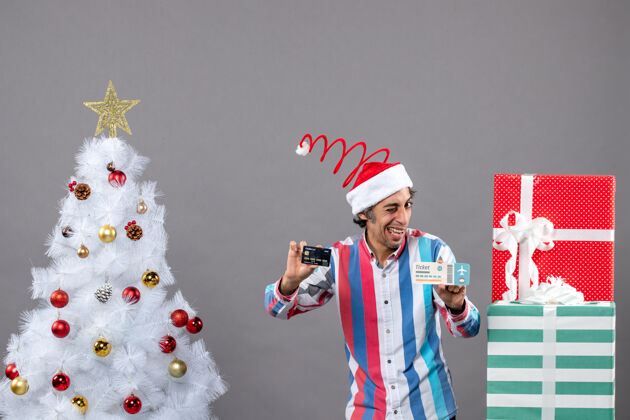 快乐的人前视图快乐的男人眨着眼睛拿着卡片和旅行票绕着圣诞树和礼物眨眼灰色人