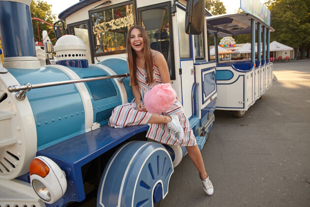 快乐阳光和煦的日子里 快乐的长发黑发女人穿着浅色夏装坐在游乐园的蒸汽火车车厢里 手里拿着棉花糖积极户外头发