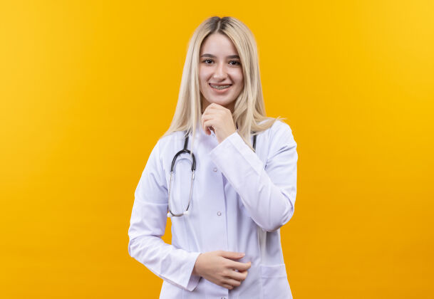 医疗微笑的医生年轻的女孩穿着医用长袍戴着听诊器和牙套把她的手放在下巴上孤立的黄色背景女孩微笑支架