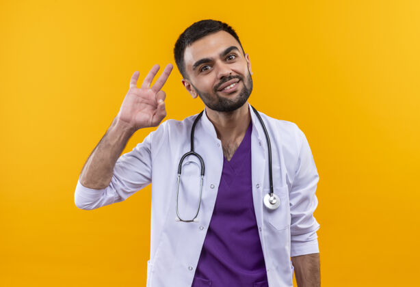 微笑微笑着的年轻男医生穿着听诊器医用长袍 在孤立的黄色背景上显示出良好的姿态手势听诊器年轻