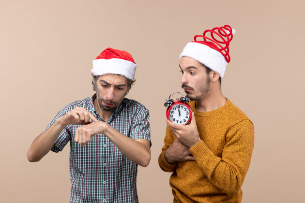 视图正面图两个男人 一个拿着闹钟 另一个在米色孤立的背景上问时间拿着帽子时间