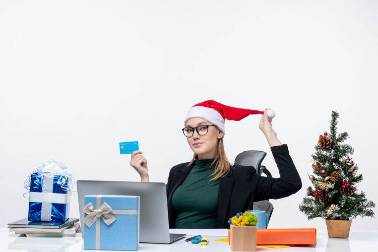 玩着一个积极迷人的女人 戴着圣诞老人的帽子 戴着眼镜 坐在桌子旁 手里拿着银行卡在办公室里正面女人桌子