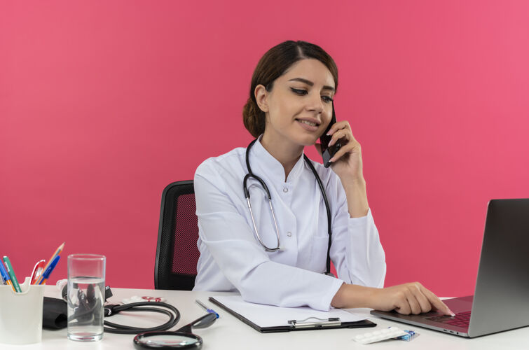 电话年轻的女医生穿着医用长袍 手持听诊器 坐在办公桌旁 拿着医疗工具在电脑上工作 在电话里讲话 用笔记本电脑复印医生高兴笔记本电脑