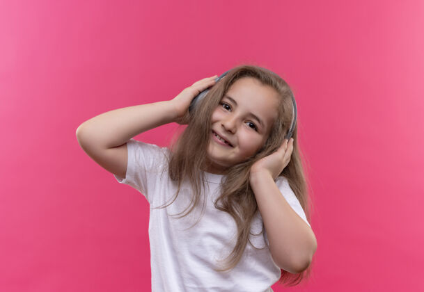 小微笑的小女生穿着白色t恤 在孤立的粉色背景下 用耳机听音乐女孩粉色T恤