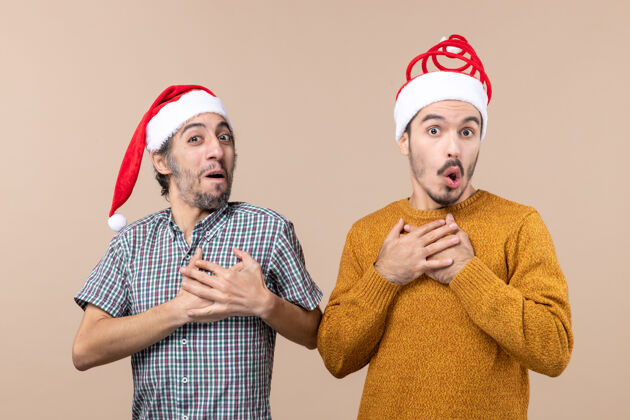 圣诞老人正面图：两个戴着圣诞帽的惊讶的家伙把手放在米色孤立背景的胸前胸部两个前面