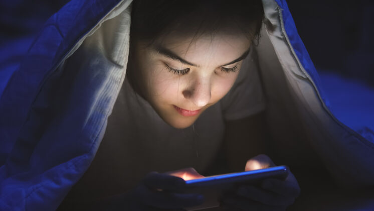 科技微笑的少女躺在床上用智能手机和浏览互联网的色调肖像移动短信女性