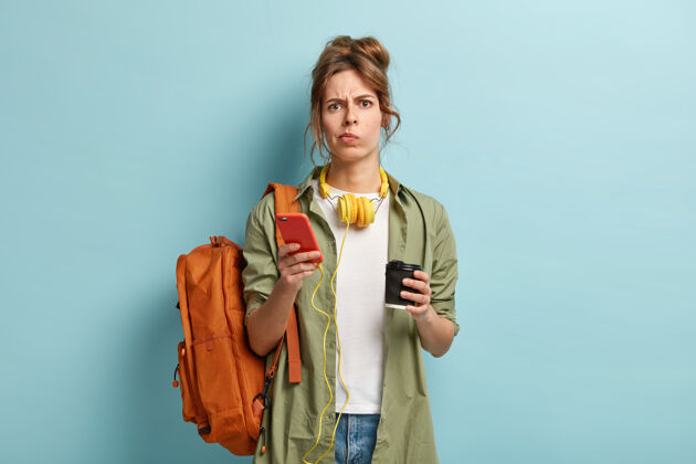 电话沮丧疲惫的千禧一代女孩喝外卖咖啡 手持智能手机设备连接到耳机 享受播放列表耳机手机皱眉