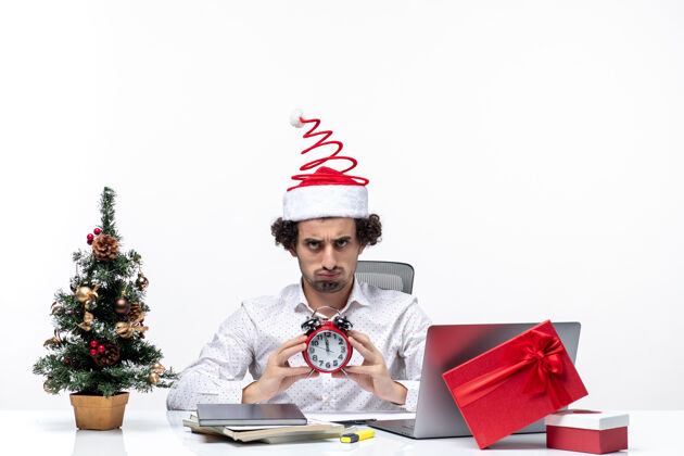 办公室戴着圣诞老人帽 拿着闹钟 满脸胡须的年轻商人惊讶地看着什么东西 坐在办公室的白色背景股票照片上无辜的惊喜商务人士