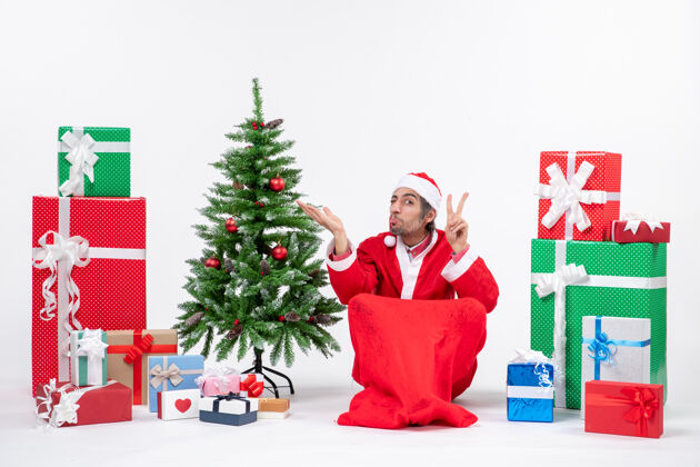 指向一脸酸酸的年轻人打扮成圣诞老人 拿着礼物和装饰好的圣诞树坐在地上做着胜利的手势 指着白色背景上的东西卖家坐着年轻人