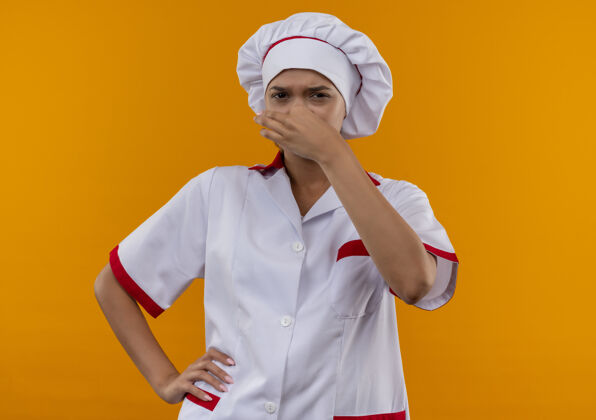 女神经质的年轻女厨师穿着厨师制服闭着鼻子把她的手放在臀部孤立的橙色背景穿厨师臀部
