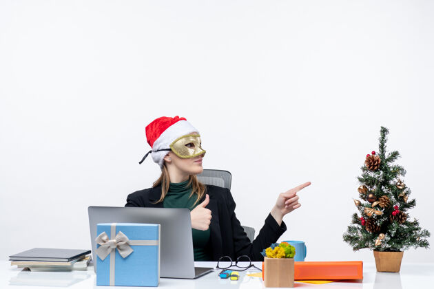 什么圣诞气氛 戴着圣诞老人帽 戴着面具的年轻女子坐在桌子旁 指着白色背景上的东西桌子面具电脑