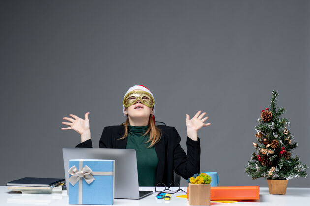 帽子圣诞气氛与戴着圣诞老人帽子和面具的年轻女子坐在一张桌子上看上面的黑暗背景圣诞老人桌子办公室