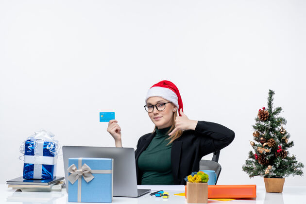 电脑自信迷人的女人 戴着圣诞老人的帽子 戴着眼镜 坐在桌子旁 手里拿着银行卡 在办公室里做着给我打电话的手势女人迷人的女人桌子