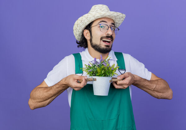 男人年轻的留着胡子的园丁 穿着连体衣 戴着帽子 手里拿着盆栽植物 高兴地笑着园丁植物站着