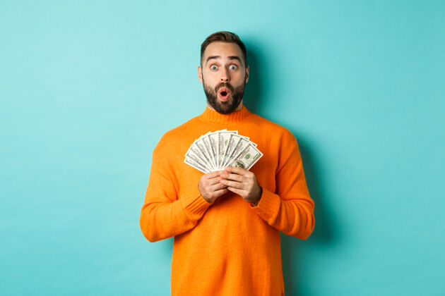毛衣照片上的惊讶的家伙拿着钱 看起来很惊讶 站在绿松石墙美元肖像年轻休闲