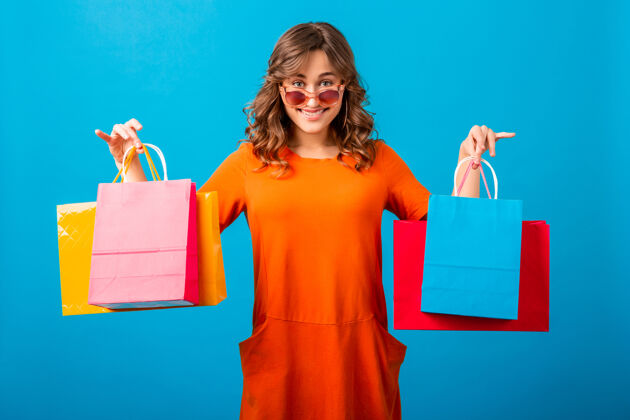 微笑迷人的快乐情绪微笑时尚的女人购物狂在橙色时尚的超大礼服举行购物袋蓝色工作室背景隔离开朗商店女性