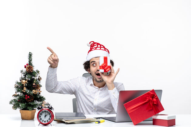 人圣诞节心情惊讶的商人与圣诞老人的帽子举起他的礼物 他的脸 并指出白色背景上的东西礼物孩子微笑