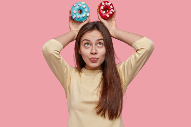 青少年一张迷人的黑发女人的照片 把五颜六色的甜甜圈放在头上 展示垃圾食品笑话零食甜点