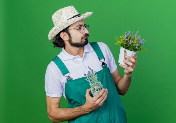帽子年轻的留着胡子的园丁 穿着连体衣 戴着帽子 手里拿着盆栽植物 严肃地看着它们园丁年轻胡须