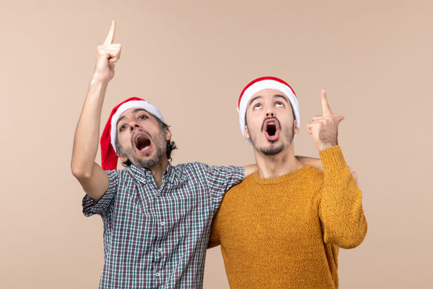 米色正面图两个热情的家伙戴着圣诞帽 在米色孤立的背景上展示了一些东西表情男人脸