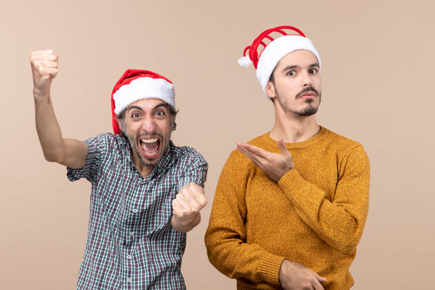 两个家伙前视图两个家伙一个兴高采烈 一个困惑的圣诞帽一个显示在米色孤立的背景其他兴高采烈圣诞老人困惑