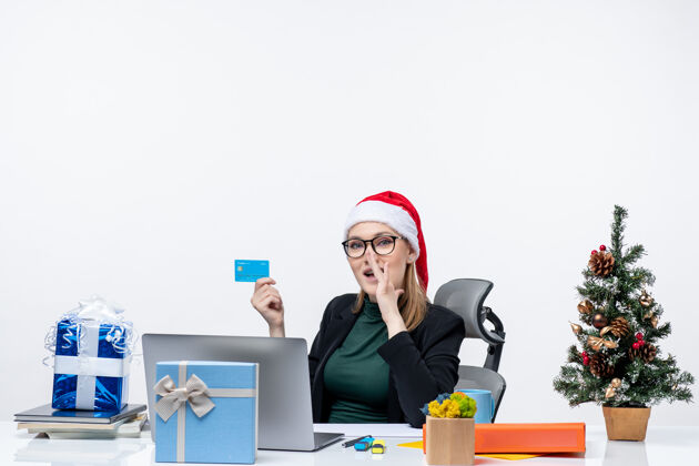帽子自信迷人的女人 戴着圣诞老人的帽子 戴着眼镜 坐在桌子旁 拿着银行卡 在办公室里闲聊抱着迷人自信