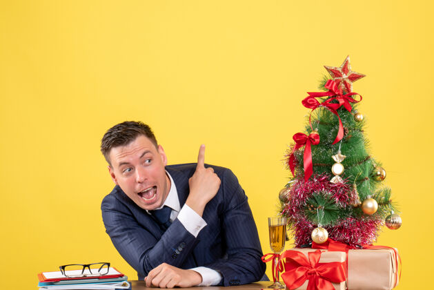 年轻人年轻人用手指着坐在圣诞树旁的桌子上 拿着黄色的礼物男人桌子手指
