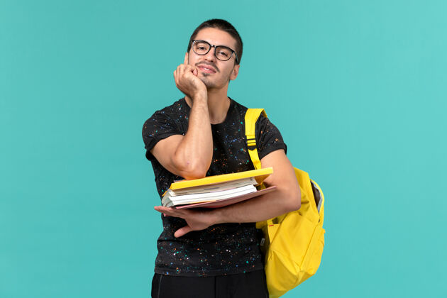 举行身穿深色t恤黄色背包的男学生正面图 浅蓝色墙上有文件和书籍男性正面大学