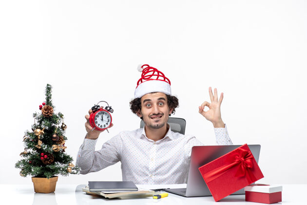 年轻人戴着圣诞老人帽 拿着钟表 戴着眼镜的年轻商务人士坐在白色背景的办公室里商务圣诞老人手势