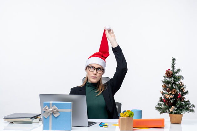 商务女人一个积极的女商人坐在一张桌子旁 手里拿着圣诞树和礼物 手里拿着圣诞老人的帽子 想着白色背景上的东西圣诞节帽子办公室