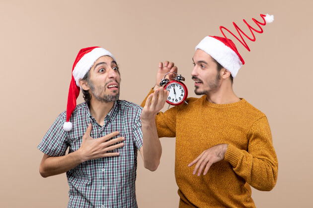 帽子前视图两个圣诞老人一个在展示什么 另一个拿着闹钟 背景是米色的男性闹钟米色
