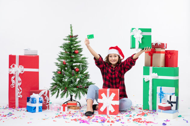 人正面图：年轻女子手持绿色银行卡和白墙上的礼物围坐在一起快乐圣诞节坐着