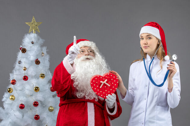 男人圣诞老人和女医生的前视图 女医生在灰色墙上用听诊器观察礼物圣诞老人圣诞医生